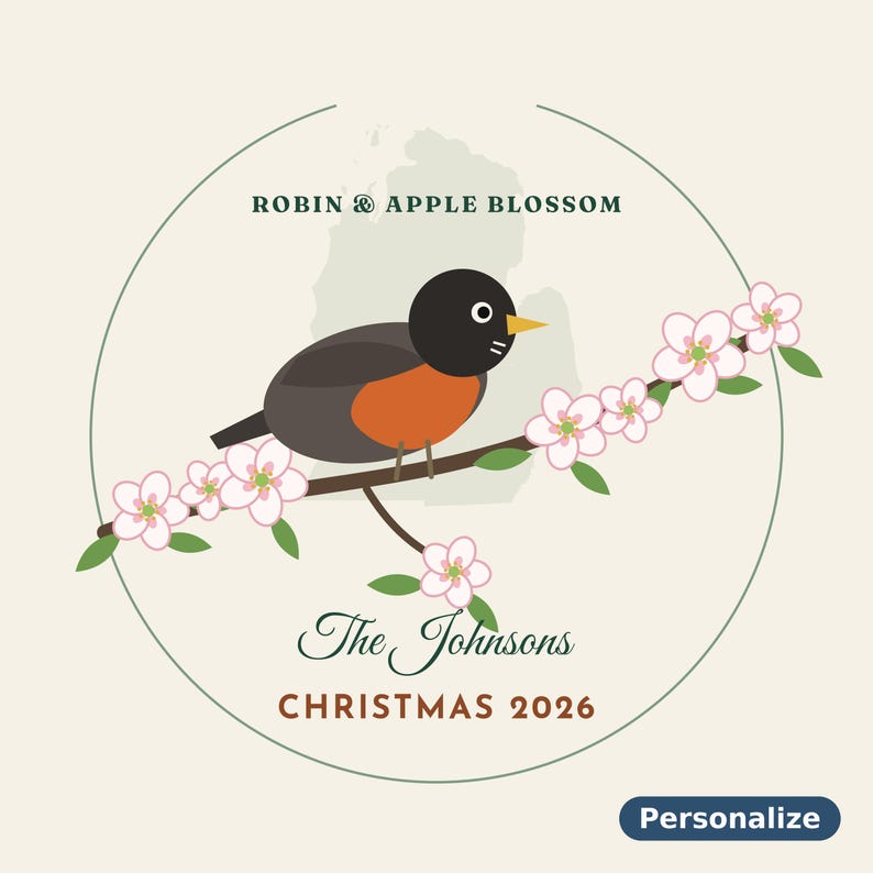 May include: A circular graphic design featuring a robin perched on an apple blossom branch. Text reads ROBIN & APPLE BLOSSOM, The Johnsons, and CHRISTMAS 2026. A faint map outline is in the background. A blue button labeled Personalize is included.