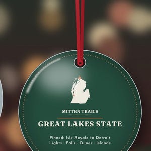 May include: A round, dark green ornament hanging from a red ribbon. The center features a white map of Michigan with a dotted line, the text "MITTEN TRAILS", "GREAT LAKES STATE", and "Pinned: Isle Royale to Detroit Lights &middot; Falls &middot; Dunes &middot; Islands".