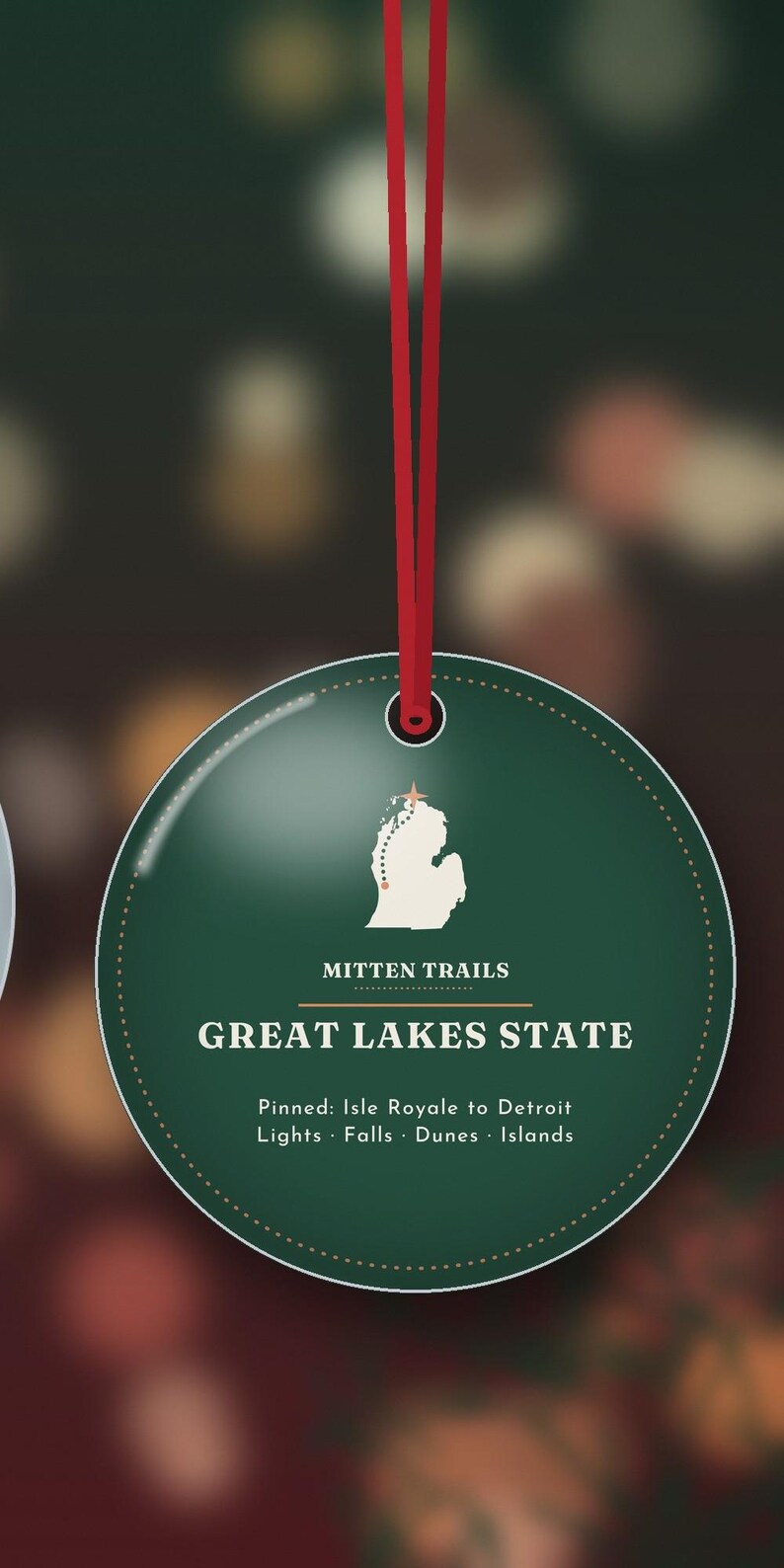 May include: A round, dark green ornament hanging from a red ribbon. The center features a white map of Michigan with a dotted line, the text "MITTEN TRAILS", "GREAT LAKES STATE", and "Pinned: Isle Royale to Detroit Lights &middot; Falls &middot; Dunes &middot; Islands".