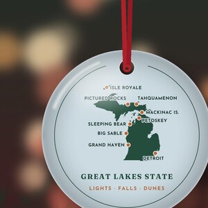 May include: A round white ornament with a red ribbon featuring a green map of Michigan. Locations marked include Isle Royale, Pictured Rocks, Tahquamenon, Mackinac Is., Petoskey, Sleeping Bear, Big Sable, Grand Haven, and Detroit. Text reads Great Lakes State, Lights, Falls, Dunes.