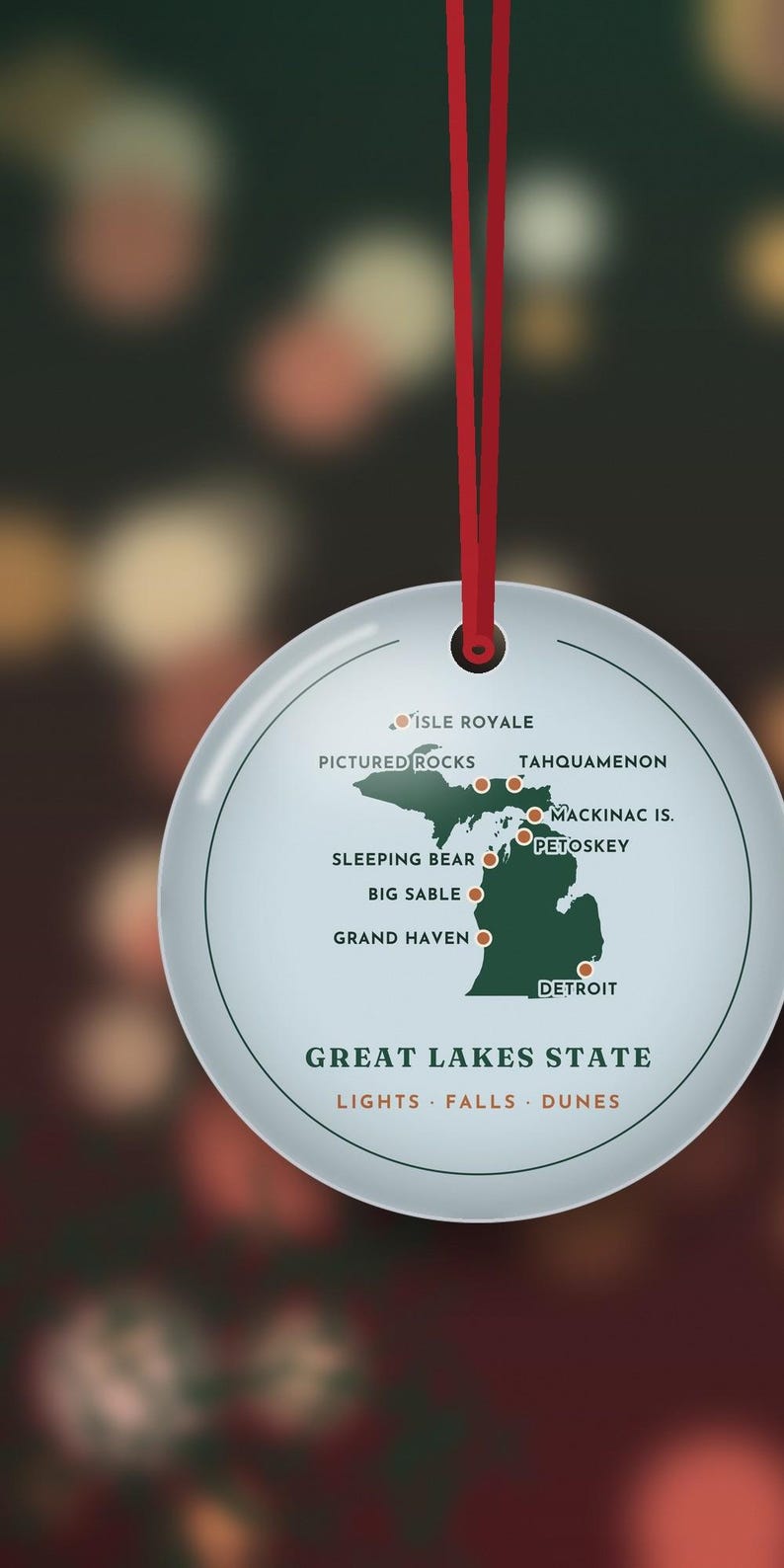 May include: A round white ornament with a red ribbon featuring a green map of Michigan. Locations marked include Isle Royale, Pictured Rocks, Tahquamenon, Mackinac Is., Petoskey, Sleeping Bear, Big Sable, Grand Haven, and Detroit. Text reads Great Lakes State, Lights, Falls, Dunes.