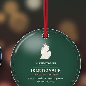 May include: A round, dark green ornament hanging from a red ribbon. It features a white map of Isle Royale, the text "MITTEN TRAILS", "ISLE ROYALE", "48&deg;00'36"N 88&deg;51'W", and "400+ islands in Lake Superior" with "Moose country" printed below.