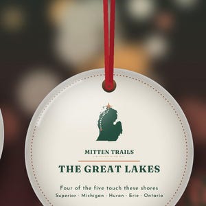 May include: A round white ornament hanging from a red ribbon. It features a green silhouette of Michigan with a red star and dotted line, the text MITTEN TRAILS, THE GREAT LAKES, and Four of the five touch these shores Superior Michigan Huron Erie Ontario.
