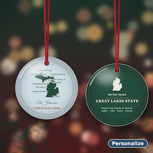 May include: Two round Christmas ornaments with red hanging strings. The left ornament is light blue with a map of Michigan, location markers, the text The Johnsons and CHRISTMAS 2026. The right ornament is dark green with a white Michigan map, MITTEN TRAILS, GREAT LAKES STATE and text.