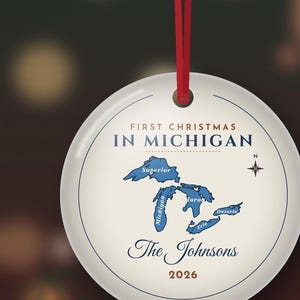 May include: A round white ceramic ornament hanging from a red ribbon. The text reads FIRST CHRISTMAS IN MICHIGAN, The Johnsons, and 2026. A blue map of the Great Lakes is centered with a compass rose. The design includes thin blue circular lines.