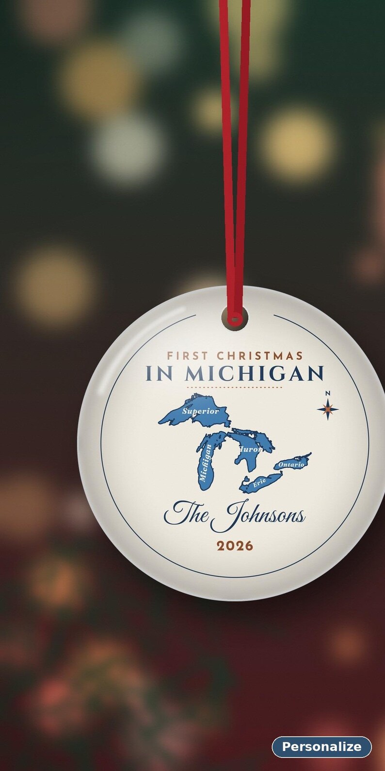 May include: A round white ceramic ornament hanging from a red ribbon. The text reads FIRST CHRISTMAS IN MICHIGAN, The Johnsons, and 2026. A blue map of the Great Lakes is centered with a compass rose. The design includes thin blue circular lines.