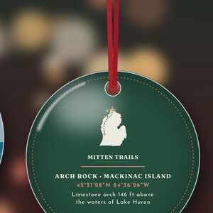 May include: A round dark green ornament hanging from a red ribbon. It features a white map of Michigan with a star, the text MITTEN TRAILS, ARCH ROCK MACKINAC ISLAND, 45&deg;51'28"N 84&deg;36'26"W, and Limestone arch 146 ft above the waters of Lake Huron.