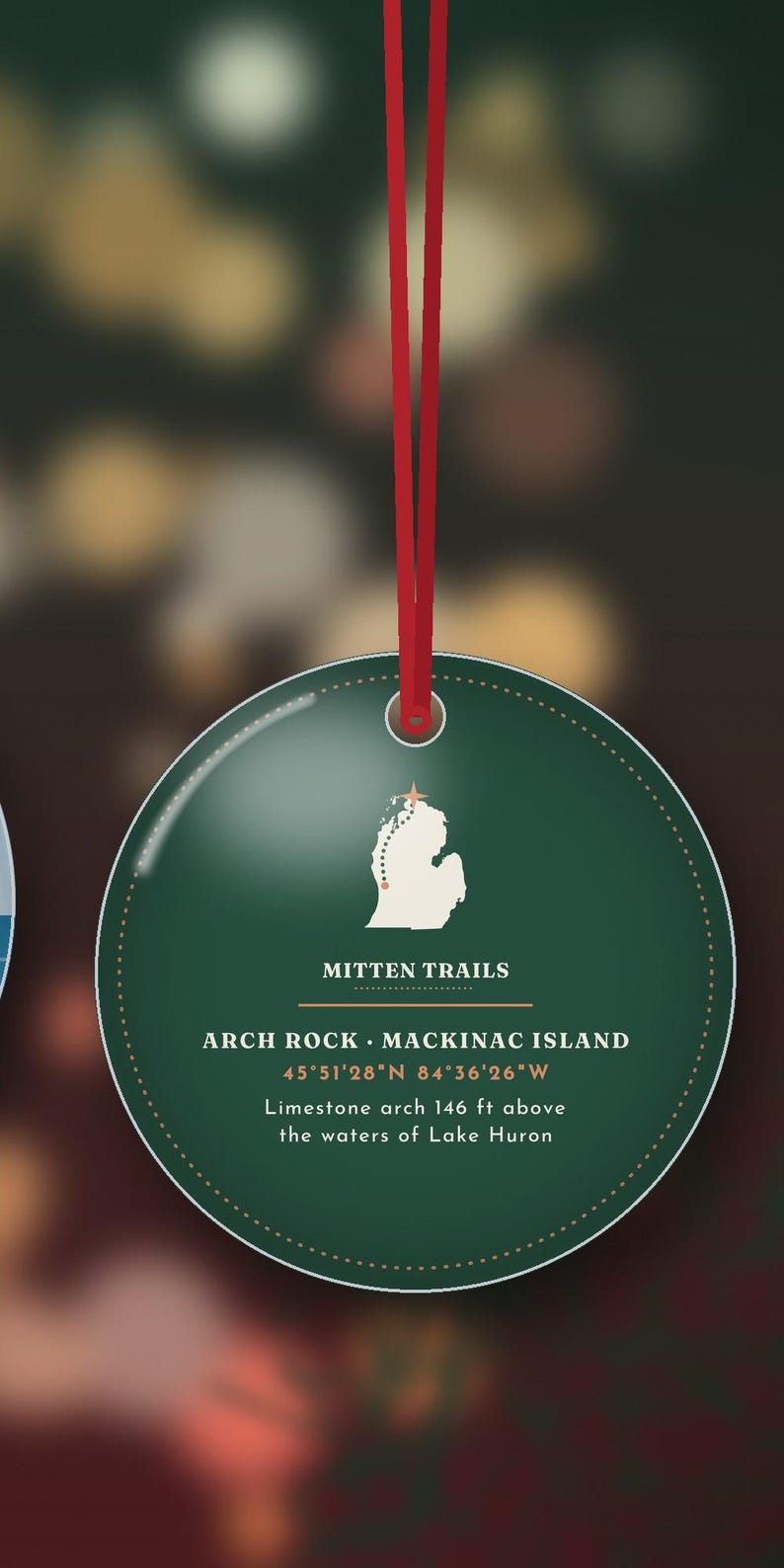 May include: A round dark green ornament hanging from a red ribbon. It features a white map of Michigan with a star, the text MITTEN TRAILS, ARCH ROCK MACKINAC ISLAND, 45&deg;51'28"N 84&deg;36'26"W, and Limestone arch 146 ft above the waters of Lake Huron.
