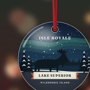 May include: A round, flat, dark blue ornament hanging from a red ribbon. The design features a silhouette of a moose in a snowy forest under a night sky with aurora borealis. Text reads: ISLE ROYALE, LAKE SUPERIOR, WILDERNESS ISLAND.