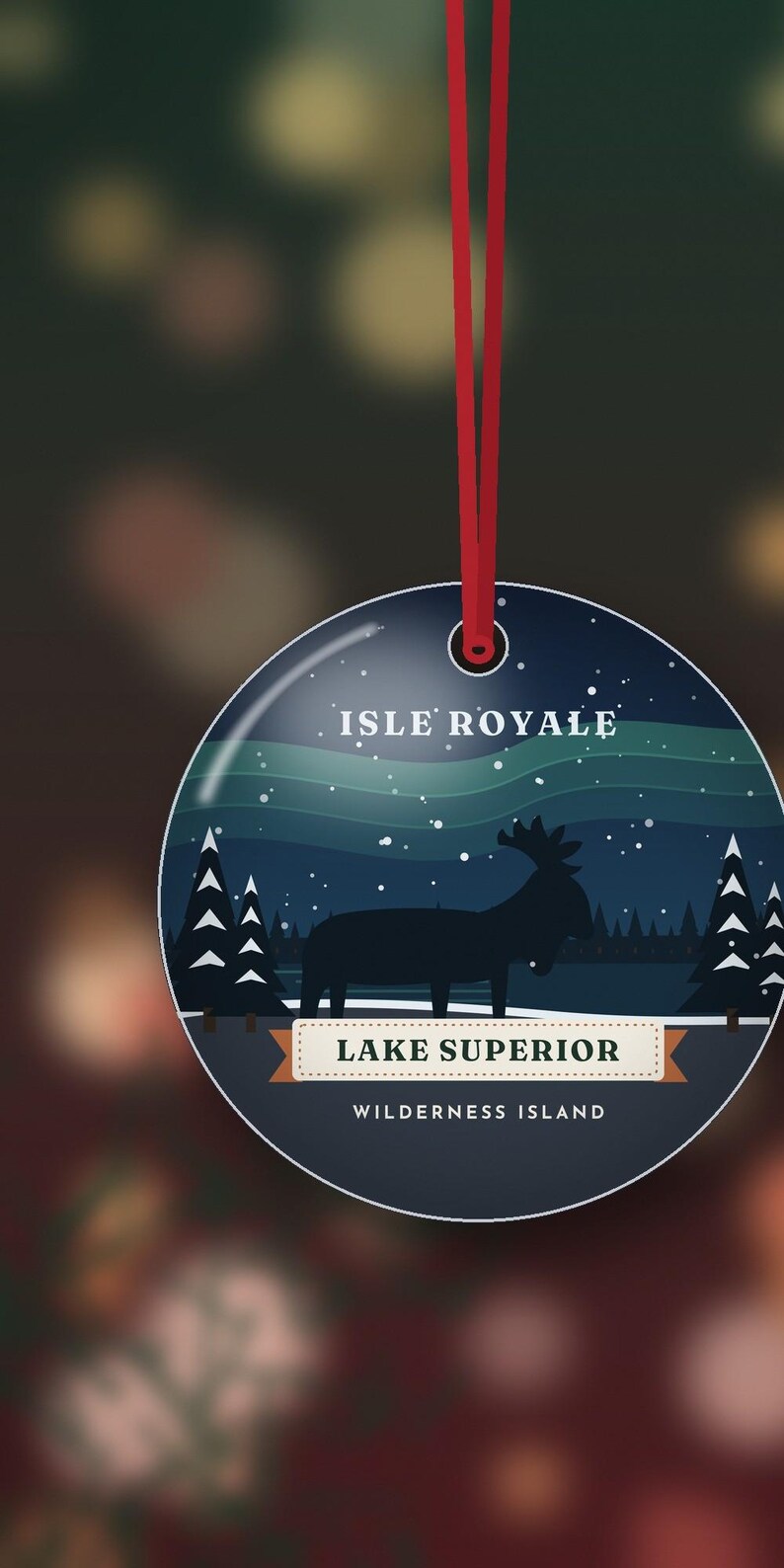 May include: A round, flat, dark blue ornament hanging from a red ribbon. The design features a silhouette of a moose in a snowy forest under a night sky with aurora borealis. Text reads: ISLE ROYALE, LAKE SUPERIOR, WILDERNESS ISLAND.