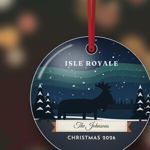May include: A round, flat, dark blue Christmas ornament hanging from a red ribbon. The design features a silhouette of a moose in a snowy forest under a green aurora borealis. Text reads ISLE ROYALE, The Johnsons, and CHRISTMAS 2026.