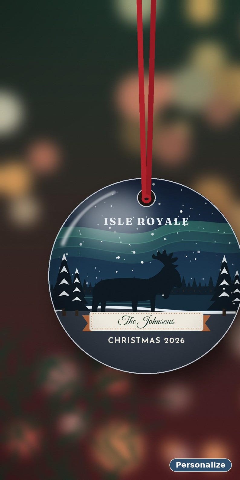 May include: A round, flat, dark blue Christmas ornament hanging from a red ribbon. The design features a silhouette of a moose in a snowy forest under a green aurora borealis. Text reads ISLE ROYALE, The Johnsons, and CHRISTMAS 2026.