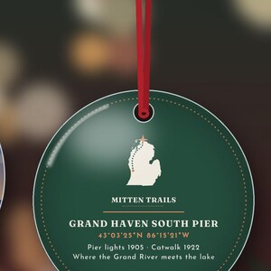 May include: A round dark green ornament hanging from a red ribbon. It features a white map of Michigan with a star, the text MITTEN TRAILS, GRAND HAVEN SOUTH PIER, coordinates 43&deg;03'25"N 86&deg;15'21"W, Pier lights 1905, Catwalk 1922, and Where the Grand River meets the lake.