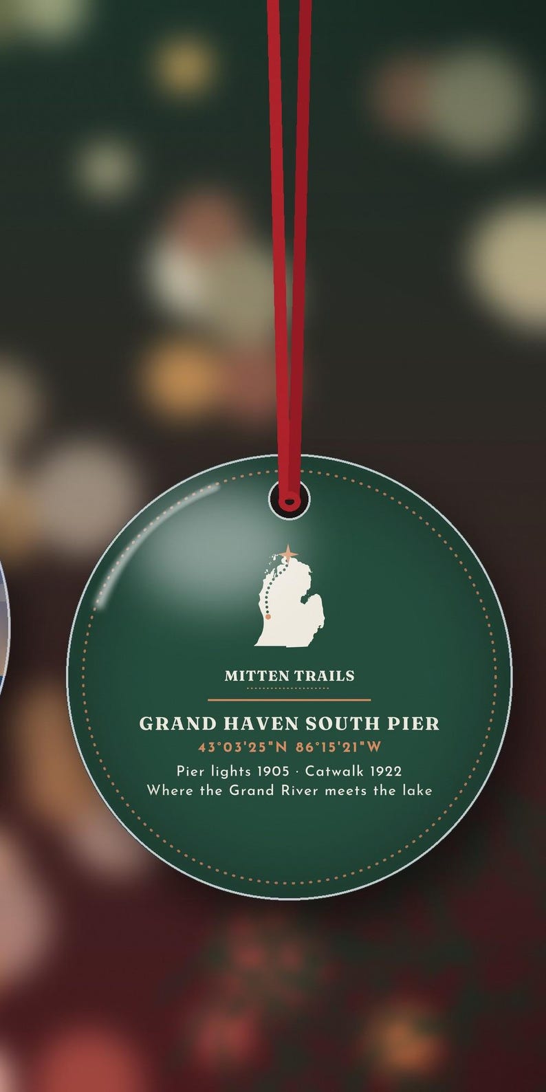 May include: A round dark green ornament hanging from a red ribbon. It features a white map of Michigan with a star, the text MITTEN TRAILS, GRAND HAVEN SOUTH PIER, coordinates 43&deg;03'25"N 86&deg;15'21"W, Pier lights 1905, Catwalk 1922, and Where the Grand River meets the lake.