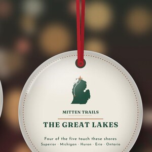 May include: A round, off-white hanging ornament with a red ribbon. It features a green silhouette of Michigan, the text "MITTEN TRAILS," "THE GREAT LAKES," and "Four of the five touch these shores Superior Michigan Huron Erie Ontario."