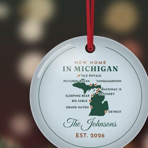 May include: A round white ceramic ornament hanging from a red ribbon. It features a green map of Michigan with red dots marking locations like Detroit, Mackinac Island, and Pictured Rocks. Text reads New Home In Michigan, The Johnsons, Est. 2026.