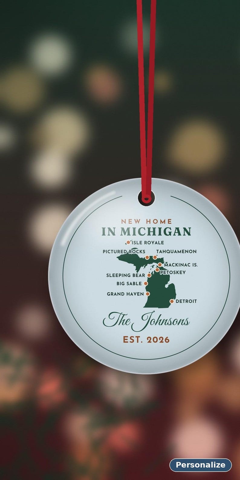 May include: A round white ceramic ornament hanging from a red ribbon. It features a green map of Michigan with red dots marking locations like Detroit, Mackinac Island, and Pictured Rocks. Text reads New Home In Michigan, The Johnsons, Est. 2026.