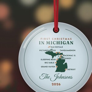 May include: A round white ceramic ornament hanging from a red ribbon. It features a green map of Michigan with red dots marking locations like Isle Royale, Pictured Rocks, and Detroit. Text reads First Christmas in Michigan, The Johnsons, and 2026.