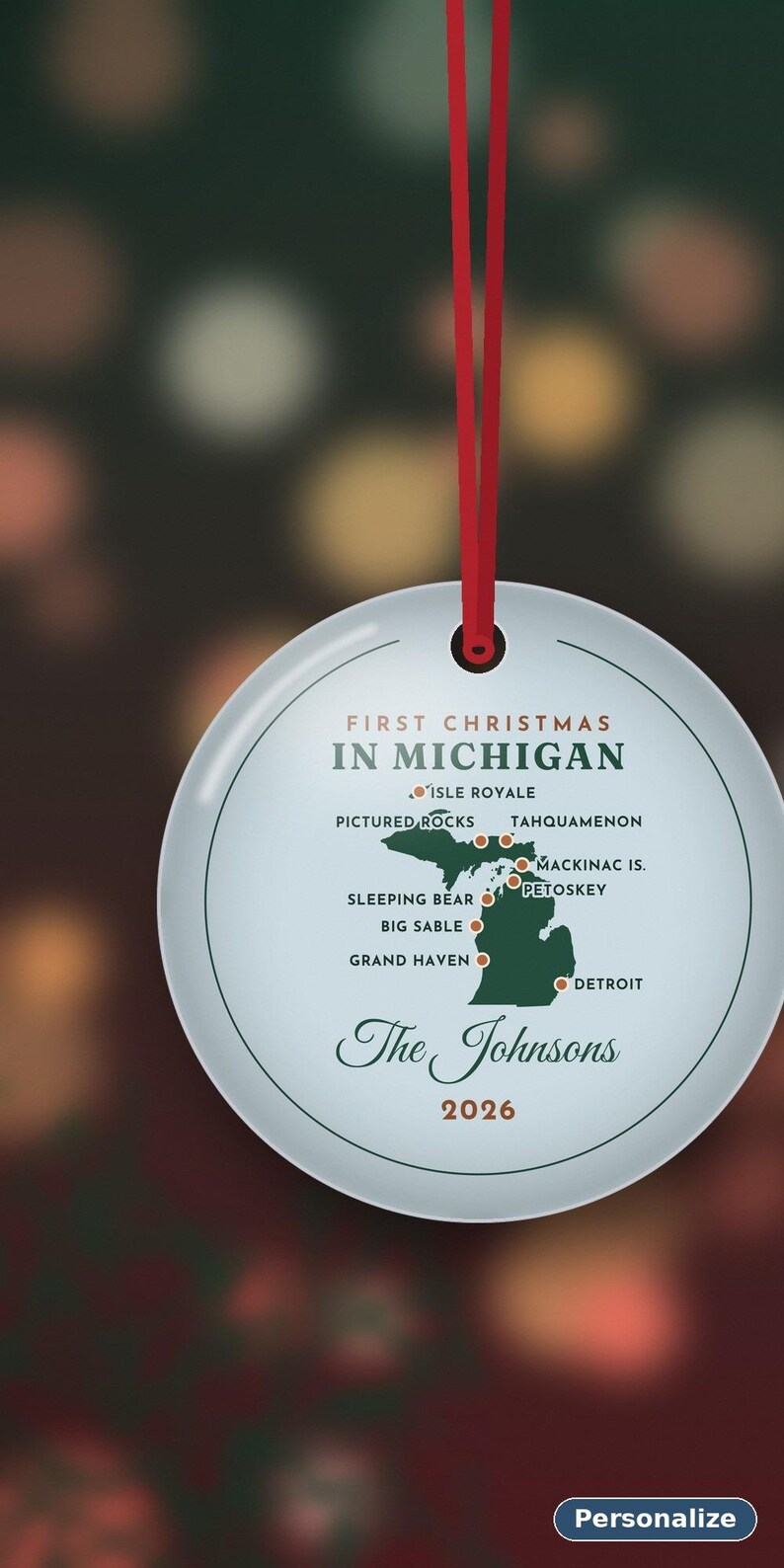 May include: A round white ceramic ornament hanging from a red ribbon. It features a green map of Michigan with red dots marking locations like Isle Royale, Pictured Rocks, and Detroit. Text reads First Christmas in Michigan, The Johnsons, and 2026.