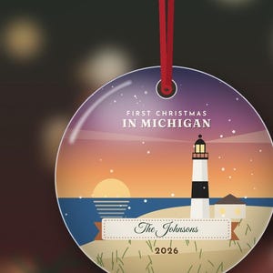 Puede incluir: Un adorno navide&ntilde;o redondo y plano que cuelga de una cinta roja. El dise&ntilde;o muestra un faro en una playa al atardecer con el texto "FIRST CHRISTMAS IN MICHIGAN", "The Johnsons" y "2026" impreso en la superficie frontal.