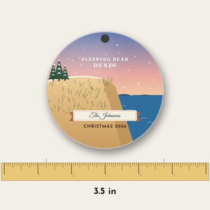 Puede incluir: Un adorno redondo con un paisaje de Sleeping Bear Dunes, una duna color canela, pinos verdes y agua azul bajo un cielo al atardecer. Incluye el texto The Johnsons y CHRISTMAS 2026. Una regla muestra un di&aacute;metro de 8,89 cm.