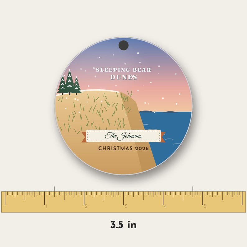 Puede incluir: Un adorno redondo con un paisaje de Sleeping Bear Dunes, una duna color canela, pinos verdes y agua azul bajo un cielo al atardecer. Incluye el texto The Johnsons y CHRISTMAS 2026. Una regla muestra un di&aacute;metro de 8,89 cm.