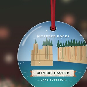 K&ouml;nnte beinhalten: Ein runder, flacher Anh&auml;nger an einer roten Schnur. Das Design zeigt einen blauen Himmel, blaues Wasser, gr&uuml;ne Tannen und beigefarbene Felsformationen. Der Text lautet: PICTURED ROCKS, MINERS CASTLE und LAKE SUPERIOR.
