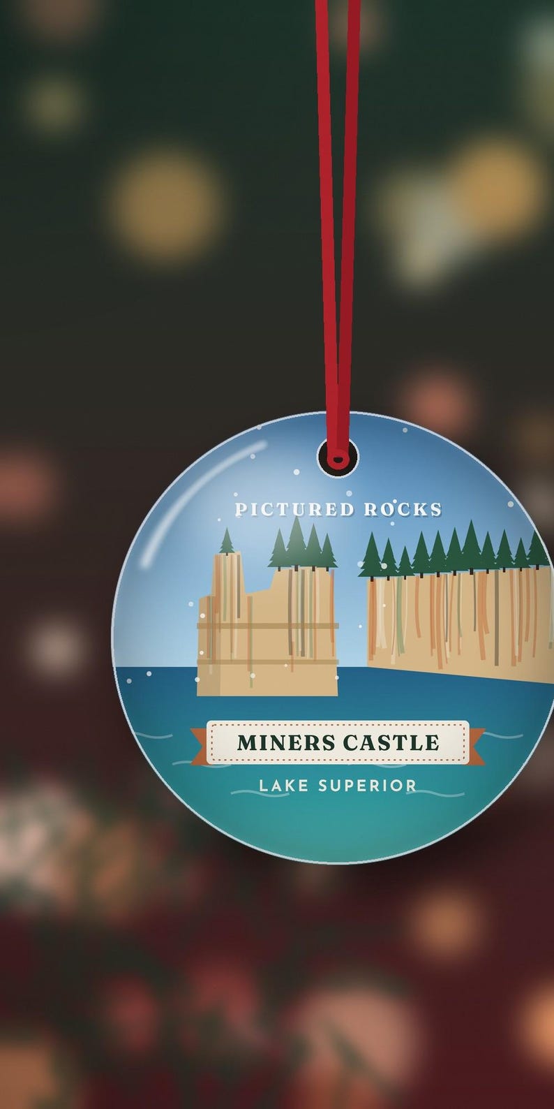 K&ouml;nnte beinhalten: Ein runder, flacher Anh&auml;nger an einer roten Schnur. Das Design zeigt einen blauen Himmel, blaues Wasser, gr&uuml;ne Tannen und beigefarbene Felsformationen. Der Text lautet: PICTURED ROCKS, MINERS CASTLE und LAKE SUPERIOR.