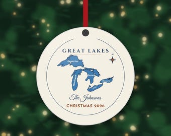 May include: A round white ceramic ornament featuring a blue map of the Great Lakes with labelled names, a compass rose, and the text "GREAT LAKES", "The Johnsons", and "CHRISTMAS 2026" in dark blue and red, suspended by a thin red ribbon.