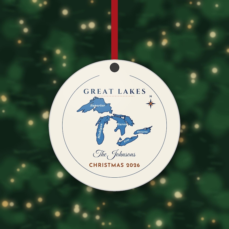 May include: A round white ceramic ornament featuring a blue map of the Great Lakes with labelled names, a compass rose, and the text "GREAT LAKES", "The Johnsons", and "CHRISTMAS 2026" in dark blue and red, suspended by a thin red ribbon.