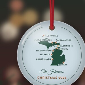 May include: A round white ceramic ornament hanging from a red ribbon. It features a green map of Michigan with orange dots marking locations like Isle Royale, Pictured Rocks, and Detroit. Text reads: The Johnsons, CHRISTMAS 2026.