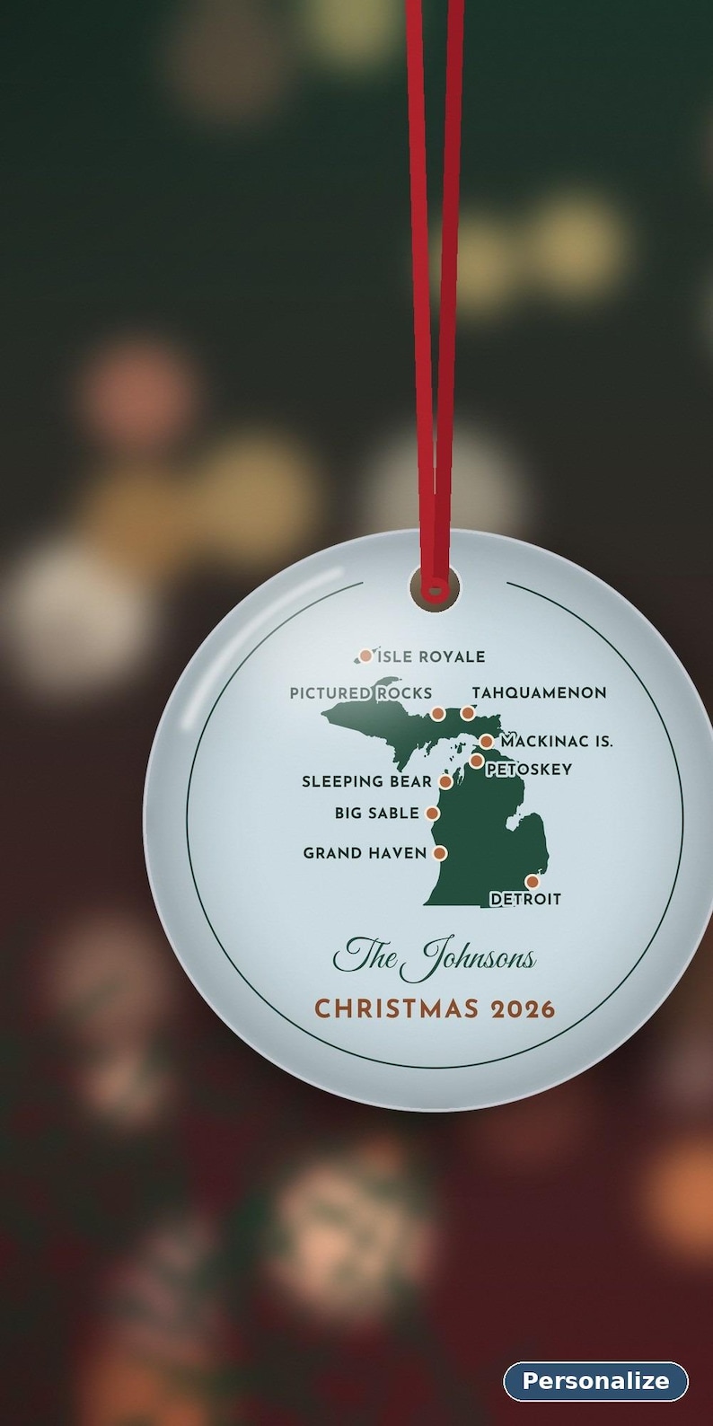May include: A round white ceramic ornament hanging from a red ribbon. It features a green map of Michigan with orange dots marking locations like Isle Royale, Pictured Rocks, and Detroit. Text reads: The Johnsons, CHRISTMAS 2026.