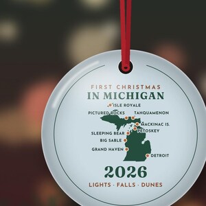 May include: A round white ceramic ornament hanging from a red ribbon. It features a green map of Michigan with red location dots and text reading FIRST CHRISTMAS IN MICHIGAN, 2026, and LIGHTS FALLS DUNES, along with specific city names.