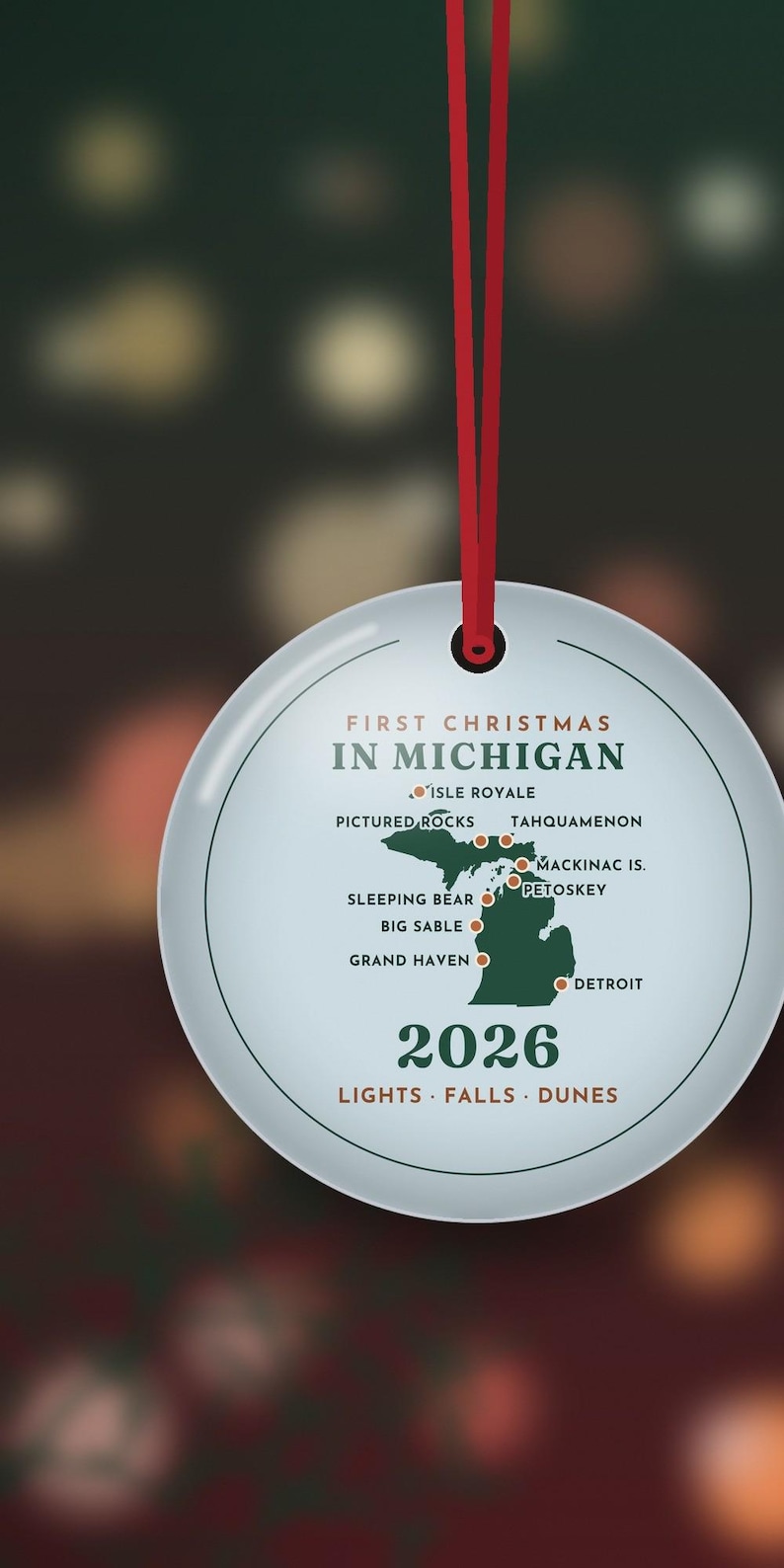 May include: A round white ceramic ornament hanging from a red ribbon. It features a green map of Michigan with red location dots and text reading FIRST CHRISTMAS IN MICHIGAN, 2026, and LIGHTS FALLS DUNES, along with specific city names.
