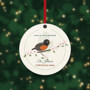 May include: A round, cream-colored ceramic ornament hanging from a red ribbon. It features a robin perched on an apple blossom branch, with the text ROBIN & APPLE BLOSSOM, The Johnsons, and CHRISTMAS 2026 printed in black and red.