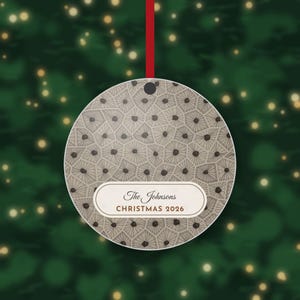 May include: A round, flat Christmas ornament featuring a beige and brown geometric pattern of interconnected polygons with dark centers. A white horizontal label displays the text The Johnsons and CHRISTMAS 2026 in brown.