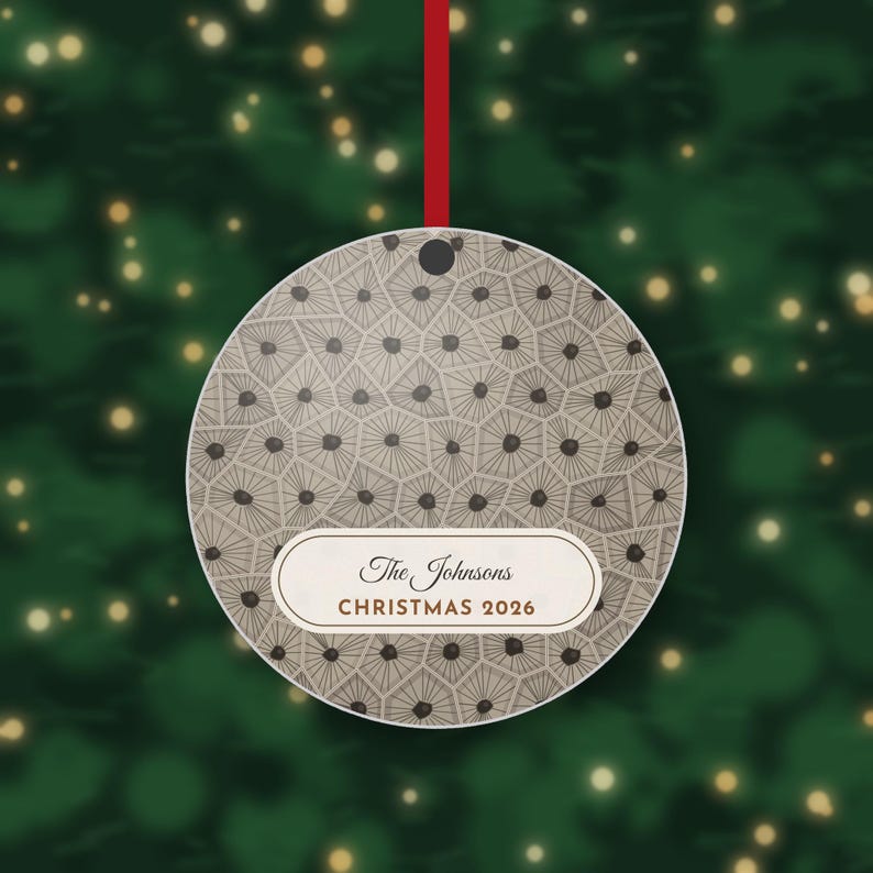 May include: A round, flat Christmas ornament featuring a beige and brown geometric pattern of interconnected polygons with dark centers. A white horizontal label displays the text The Johnsons and CHRISTMAS 2026 in brown.