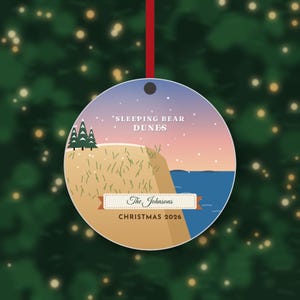 Puede incluir: Un adorno redondo de cer&aacute;mica colgado de una cinta roja, con una ilustraci&oacute;n gr&aacute;fica de Sleeping Bear Dunes con pinos verdes, arena color canela y agua azul bajo un cielo al atardecer. El texto dice Sleeping Bear Dunes, The Johnsons y Christmas 2026.