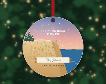 May include: A round ceramic ornament hanging from a red ribbon, featuring a graphic illustration of Sleeping Bear Dunes with green pine trees, tan sand, and blue water under a sunset sky. Text reads Sleeping Bear Dunes, The Johnsons, and Christmas 2026.