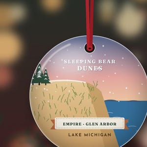 Puede incluir: Un adorno redondo y plano que cuelga de una cinta roja, con una ilustraci&oacute;n gr&aacute;fica de Sleeping Bear Dunes con un acantilado de arena color canela, pinos verdes y agua azul. El texto dice: Sleeping Bear Dunes, Empire, Glen Arbor, Lake Michigan.