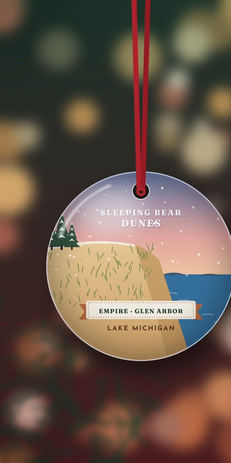 Puede incluir: Un adorno redondo y plano que cuelga de una cinta roja, con una ilustraci&oacute;n gr&aacute;fica de Sleeping Bear Dunes con un acantilado de arena color canela, pinos verdes y agua azul. El texto dice: Sleeping Bear Dunes, Empire, Glen Arbor, Lake Michigan.