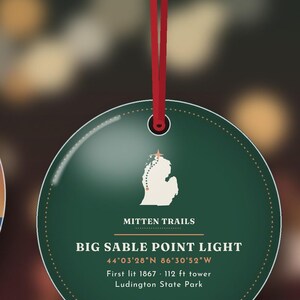K&ouml;nnte beinhalten: Ein runder, dunkelgr&uuml;ner Anh&auml;nger an einem roten Band. Er zeigt eine wei&szlig;e Karte von Michigan mit einem Stern, den Text MITTEN TRAILS, BIG SABLE POINT LIGHT, 44&deg;03'28"N 86&deg;30'52"W, First lit 1867, 34 m tower und Ludington State Park.
