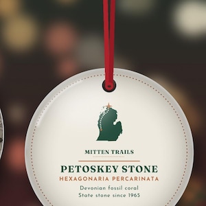 May include: A round, cream-colored ornament hanging from a red ribbon. It features a green silhouette of Michigan with a star, the text MITTEN TRAILS, PETOSKEY STONE, HEXAGONARIA PERCARINATA, Devonian fossil coral, and State stone since 1965.