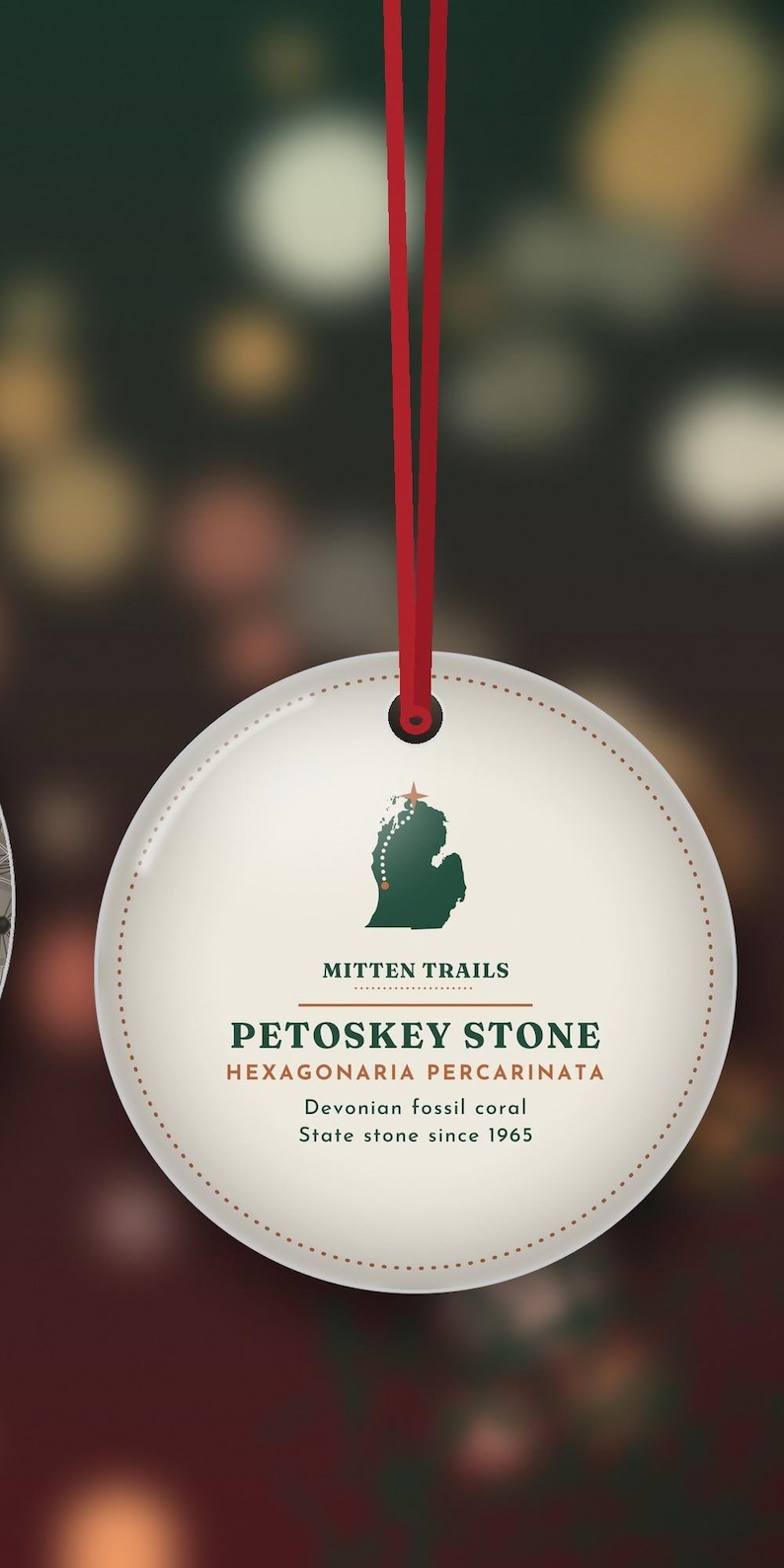 May include: A round, cream-colored ornament hanging from a red ribbon. It features a green silhouette of Michigan with a star, the text MITTEN TRAILS, PETOSKEY STONE, HEXAGONARIA PERCARINATA, Devonian fossil coral, and State stone since 1965.