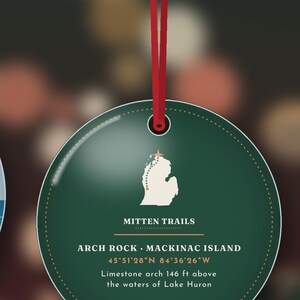 May include: A round dark green ornament hanging from a red ribbon. The centre features a white map of Michigan with a star, the text MITTEN TRAILS, ARCH ROCK MACKINAC ISLAND, 45 51'28 N 84 36'26 W, and Limestone arch 146 ft above the waters of Lake Huron.