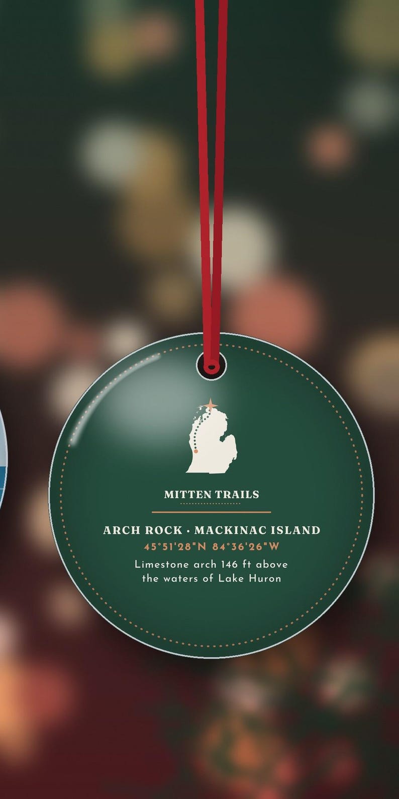 May include: A round dark green ornament hanging from a red ribbon. The centre features a white map of Michigan with a star, the text MITTEN TRAILS, ARCH ROCK MACKINAC ISLAND, 45 51'28 N 84 36'26 W, and Limestone arch 146 ft above the waters of Lake Huron.