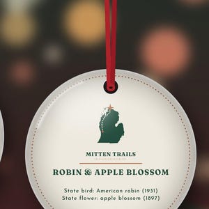 May include: A round, cream-colored hanging ornament with a red string. It features a green map of Michigan, the text 'MITTEN TRAILS', 'ROBIN & APPLE BLOSSOM', and 'State bird: American robin (1931) State flower: apple blossom (1897)'.