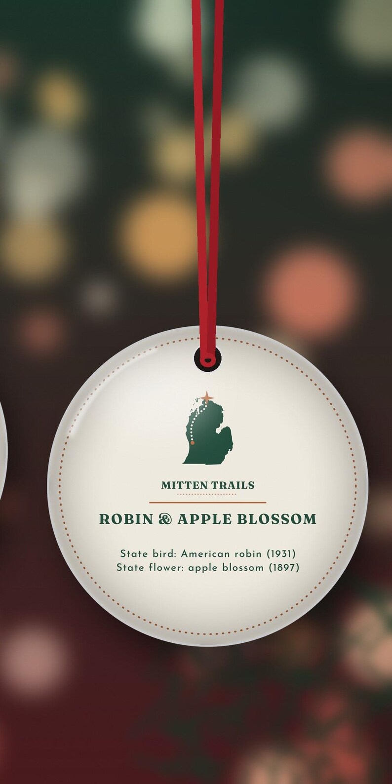 May include: A round, cream-colored hanging ornament with a red string. It features a green map of Michigan, the text 'MITTEN TRAILS', 'ROBIN & APPLE BLOSSOM', and 'State bird: American robin (1931) State flower: apple blossom (1897)'.