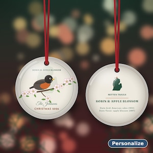 May include: Two round white ceramic ornaments with red ribbon hangers. The left ornament features a robin on an apple blossom branch, text reading ROBIN & APPLE BLOSSOM, The Johnsons, and CHRISTMAS 2026. The right shows a Michigan map, MITTEN TRAILS, and text.