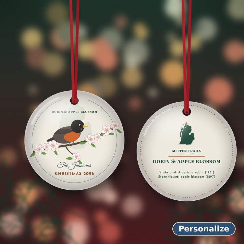 May include: Two round white ceramic ornaments with red ribbon hangers. The left ornament features a robin on an apple blossom branch, text reading ROBIN & APPLE BLOSSOM, The Johnsons, and CHRISTMAS 2026. The right shows a Michigan map, MITTEN TRAILS, and text.